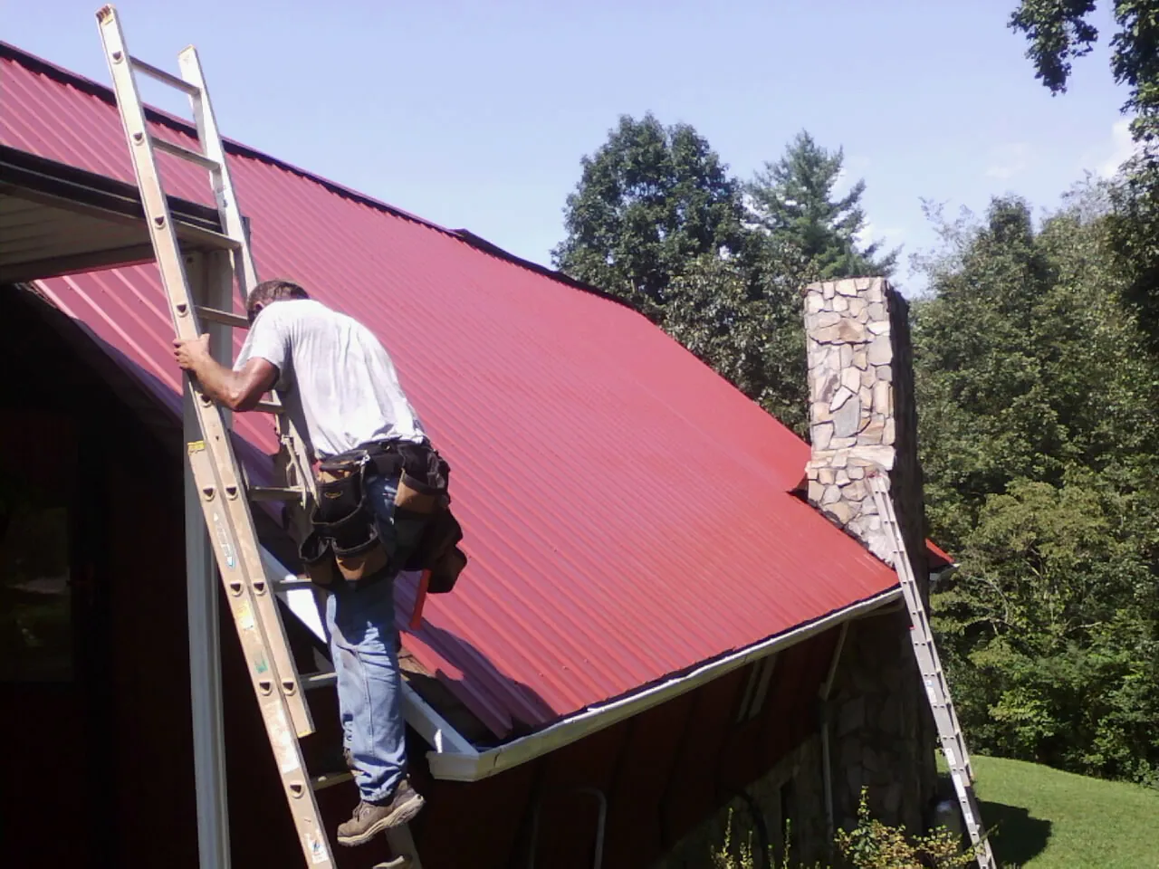 Roofer on a ladder beside a red metal roof and stone chimney — Soffit & Fascia Repair by Roofing Brothers in Emmett